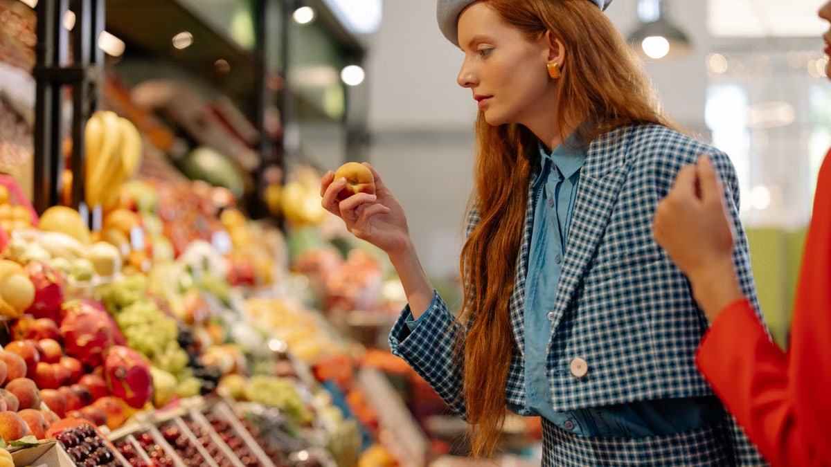 A stylish woman in a checkered blazer and beret shopping for fresh fruits at a lively market.